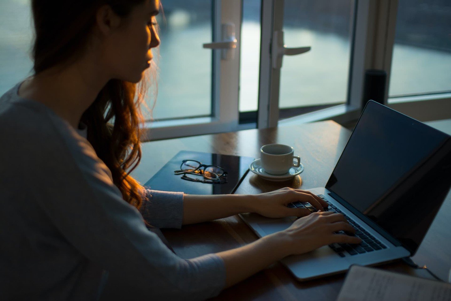 Young woman with wavy hair typing on laptop with coffee, glasses and notebook on a table Young woman with wavy hair typing on laptop with coffee, glasses and notebook on a table
